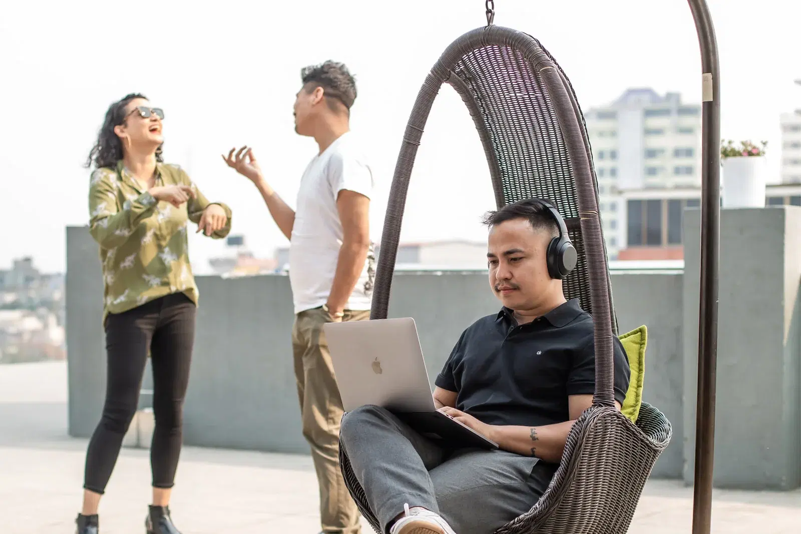 Employee on a swing chair with a laptop, two colleagues relishing the sunny day in an outdoor setting.